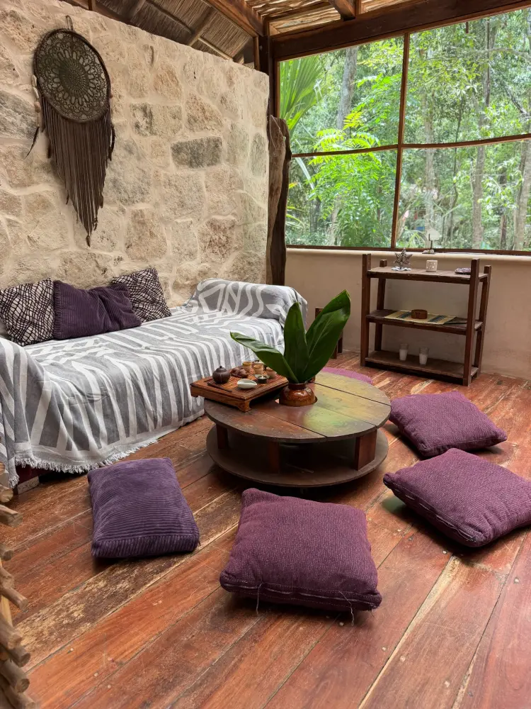 A rustic lounge with a striped sofa, wooden coffee table, and a window open to the forest